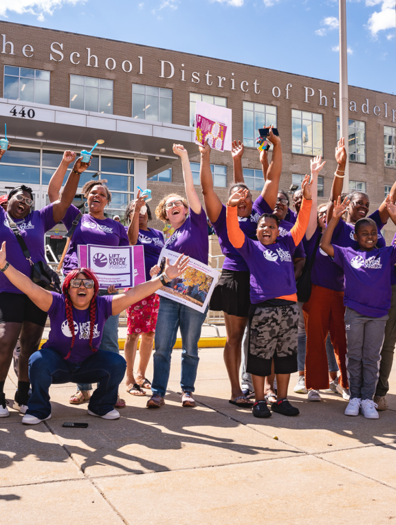 Students and adults in purple "Life Every Voice" shirts cheering in front of a school