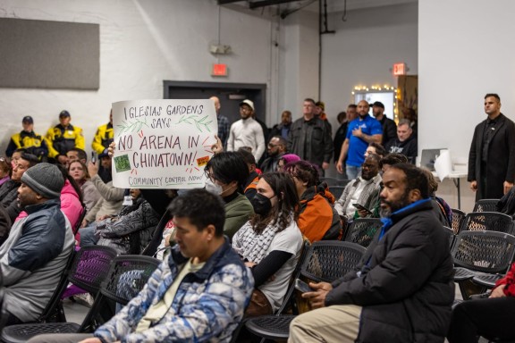 Community members listening to mayor at a town hall meeting. One community member holds a sign opposing an arena being built in Chinatown
